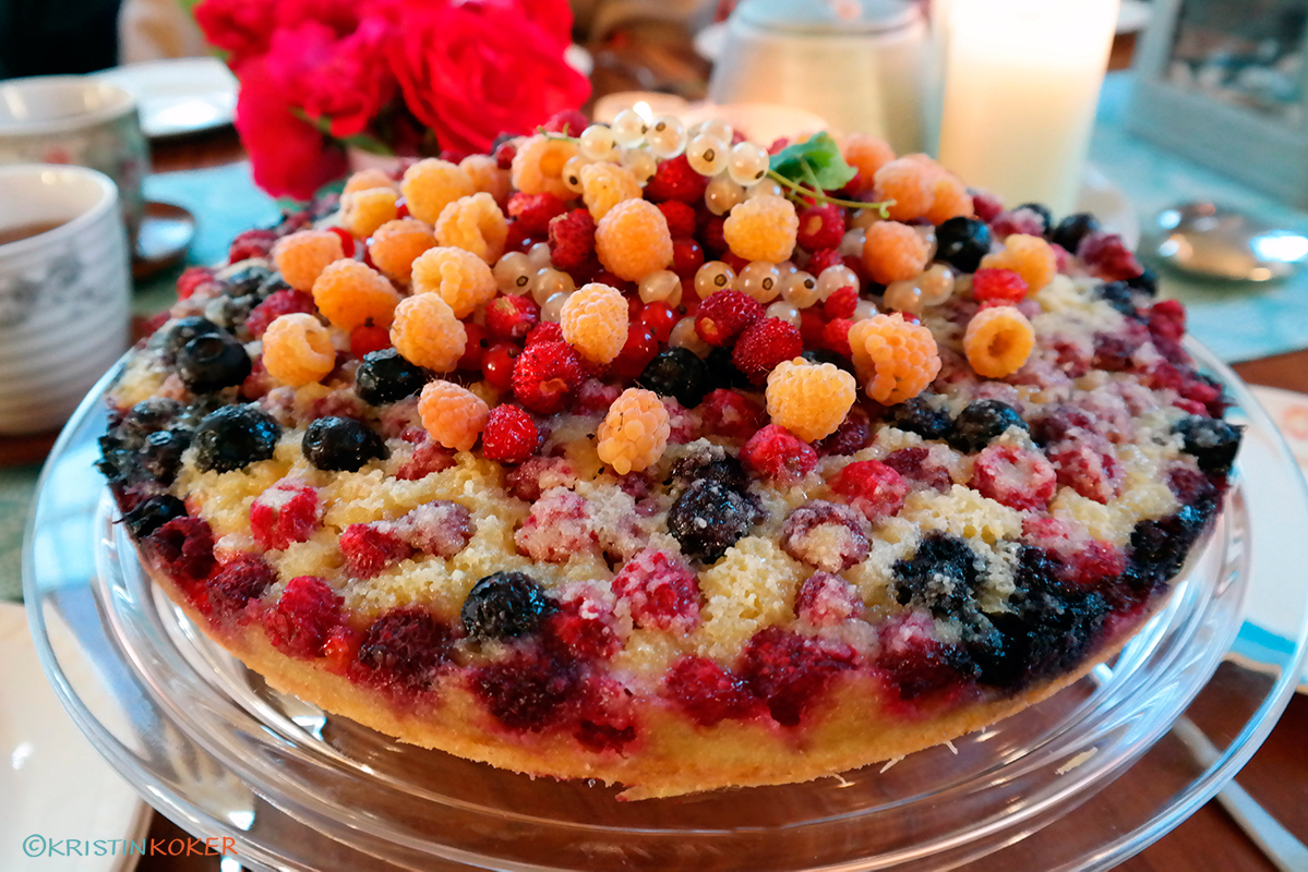 Lys kake med sommerbær og frangipanetopp. Servert på stettefat med blomster og lys i bakgrunnen.